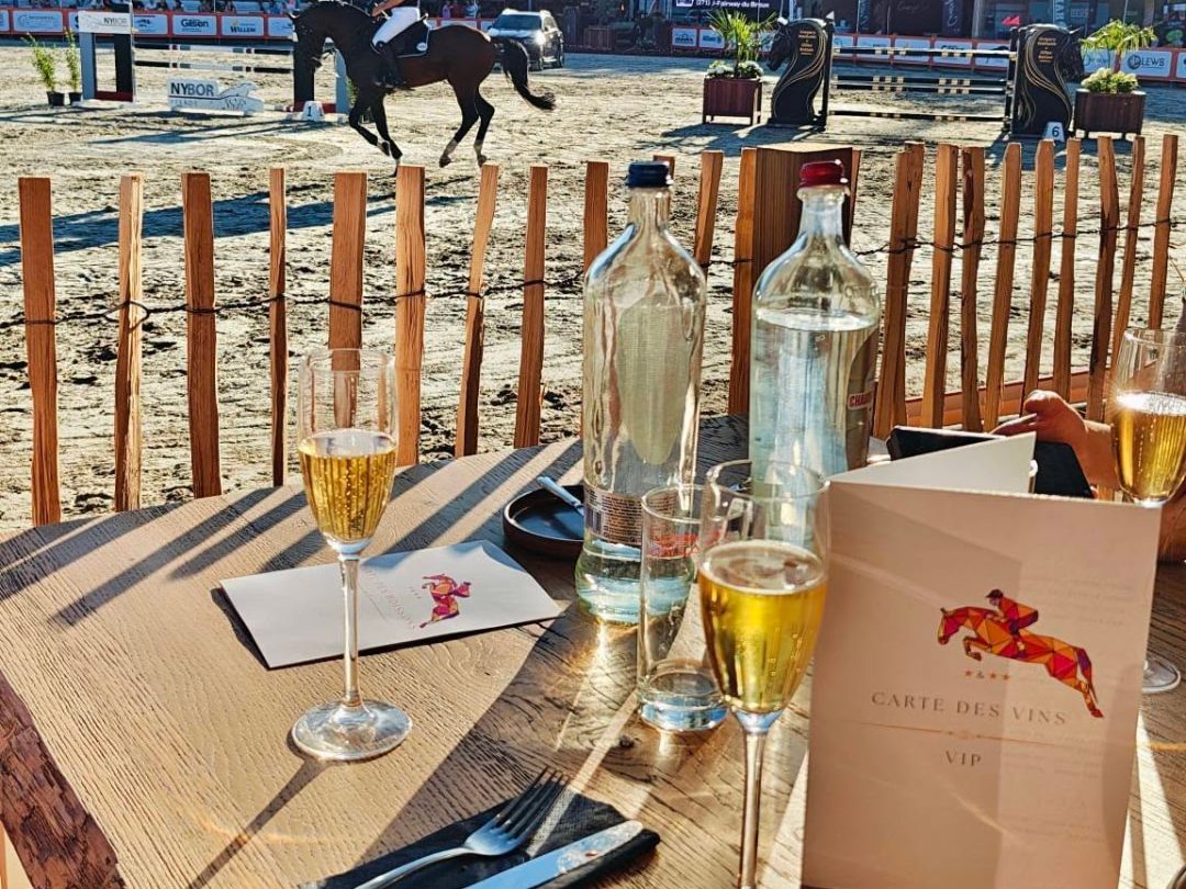 Table VIP d'EquisFair au Jumping International de Namur : table dressée avec coupes de champagne et carte des vins face à la piste d'obstacles